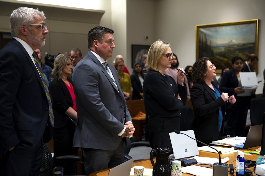 caption: Jeffrey Nelson, center, stands during his trial between his defense attorneys Tim Leary, left, and Emma Scanlan, right, as King County Superior Court Judge Nicole Gaines Phelps enters the courtroom on Wednesday, May 22, 2024, at the Maleng Regional Justice Center in Kent.