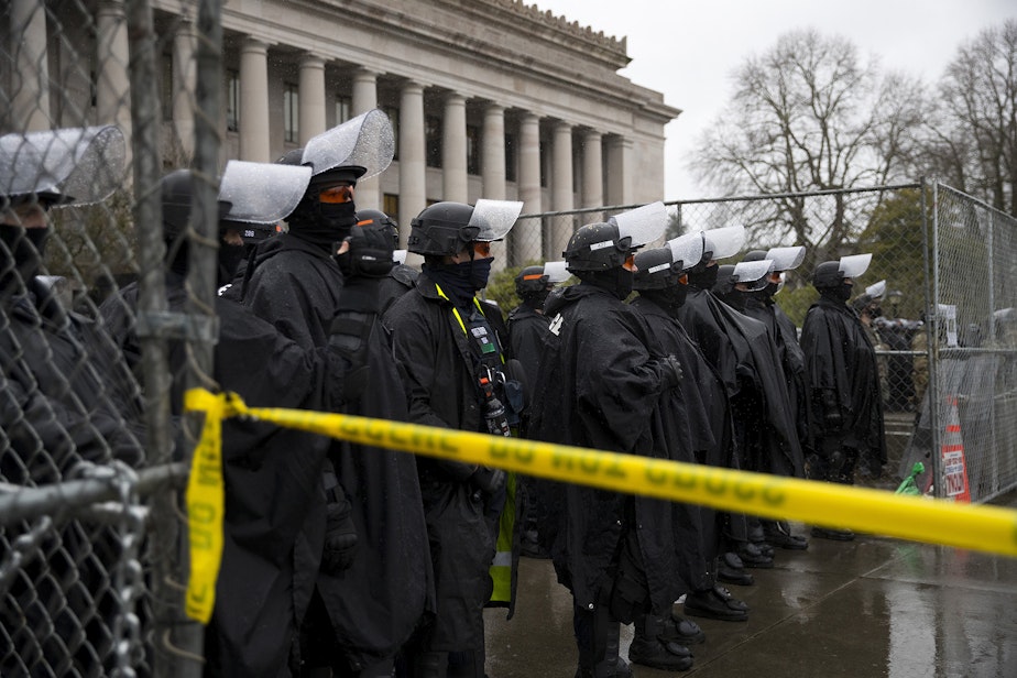 caption: Police guard the Washington State Capitol building on Monday, January 11, 2021, in Olympia. Armed protesters gathered on the opposite side of the fence on the first day of the legislative session in Olympia.