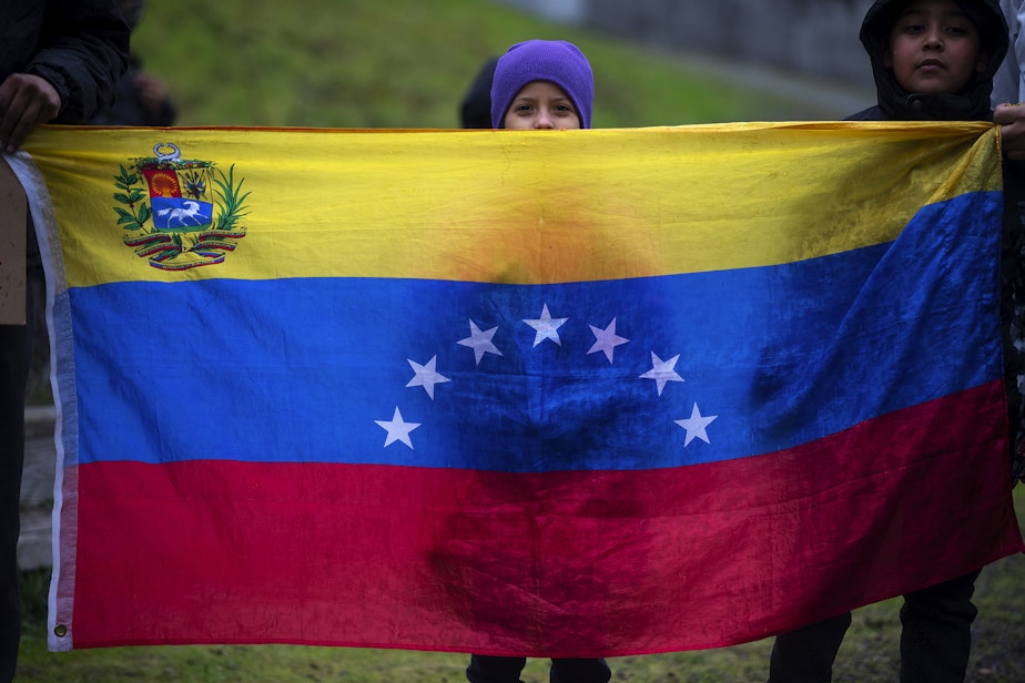 caption: A child stands behind a Venezuelan flag as asylum seekers once housed at the Sleep Inn in SeaTac, prepare to march to Seattle City Hall along with activists, mutual aid organizations and allies to ask for assistance with housing from Seattle city councilmembers on Tuesday, February 27, 2024, in Seattle.